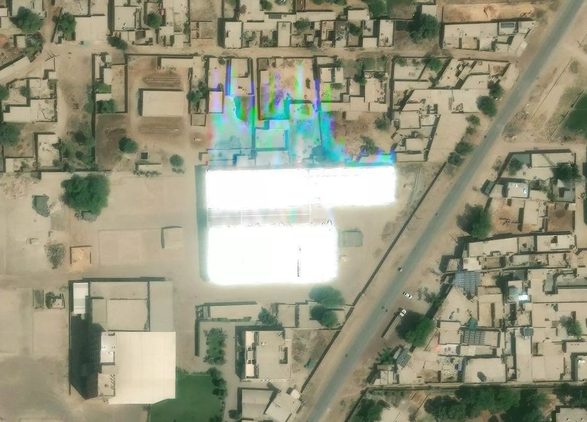 High-resolution basemap imagery of a rooftop PV array caught mid-glint: the panels saturate fully white and the overload spills off the roof as a rainbow smear of detector-blooming artifacts across the neighbouring buildings.